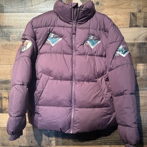 May include: A mauve puffer jacket with a high collar and a full-length front zipper. The jacket features embroidered patches on the chest and sleeves, including the text Mighty Ducks Anaheim, and has ribbed cuffs at the ends of the long sleeves.