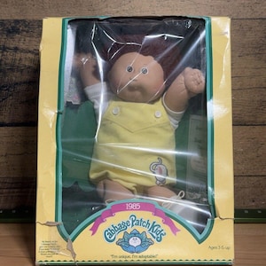 Puede incluir: Una muñeca Cabbage Patch Kids de 1985 con cabello castaño, vistiendo una camisa blanca y un overol amarillo con un gráfico de elefante. La muñeca está dentro de una caja de cartón amarilla y verde con el texto Cabbage Patch Kids, 1985 e I'm unique. I'm adoptable!