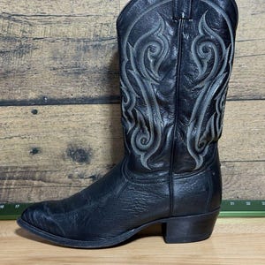 May include: A black leather cowboy boot featuring a textured finish and a tall shaft adorned with intricate grey decorative stitching. The boot has a pointed toe, a stacked heel, and pull tabs at the top of the shaft for ease of wear.