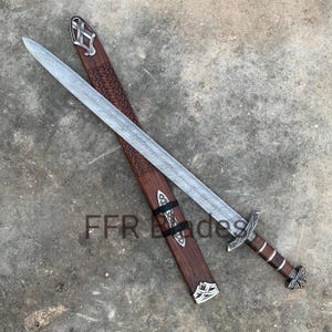 May include: A Damascus steel sword with a patterned blade, a silver-toned crossguard, and a brown leather-wrapped grip. The matching wooden scabbard features intricate carved knotwork, silver-toned metal accents, and the text FFR Blades.