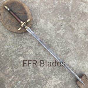 May include: A sword with a patterned steel blade, a brass crossguard, and a dark brown cylindrical handle with a metallic pommel. The text FFR Blades is visible on the blade. The sword rests diagonally across a circular wooden base.