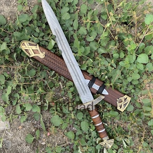 May include: A double-edged sword with a patterned steel blade, a brown leather-wrapped handle, and brass-colored metal accents. The sword is accompanied by a matching brown wooden scabbard featuring intricate carvings and brass-colored metal fittings.