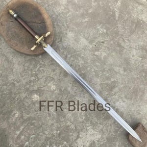 May include: A sword with a long, double-edged steel blade, a dark brown cylindrical grip, and a brass crossguard featuring decorative detailing. The pommel is rounded and metallic, and the text FFR Blades is visible on the blade.