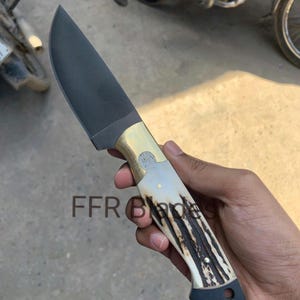 May include: A fixed-blade knife with a dark matte finish on the blade, a polished brass bolster, and a textured antler handle with brass pins. The handle features a dark pommel with a lanyard hole. The text FFR BLADES is visible on the blade.