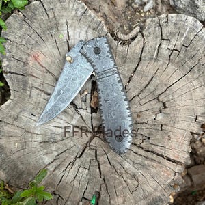 May include: A folding pocket knife featuring a distinctive grey and black Damascus steel pattern on both the blade and the handle. The blade includes a small brass thumb stud for opening, and the handle is secured with black hardware screws.