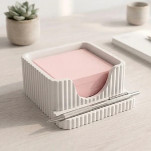May include: A white square desk organiser with a ribbed exterior texture, containing a stack of light pink square sticky notes. A silver metallic pen rests horizontally in a dedicated groove along the front edge of the organiser base.