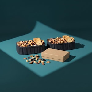 May include: Two dark gray, rectangular, ribbed containers filled with a mixture of assorted nuts, dried fruit, and square crackers. A small, plain, light brown cardboard box sits in the center, with a scattering of loose nuts and dried fruit nearby.