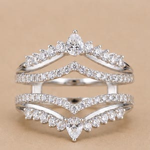 May include: A silver-toned ring guard featuring two chevron-shaped bands connected by vertical bars. The bands are encrusted with numerous small round clear stones and each features a single pear-shaped clear stone at the center point.