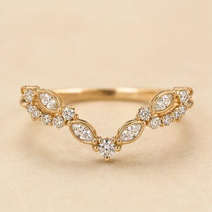May include: A gold V-shaped ring featuring a curved band set with multiple clear, sparkling stones. The design includes a mix of marquise-cut and round-cut stones arranged in a symmetrical pattern that follows the contour of the band.