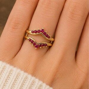 Peut inclure: Une bague double en or compos&eacute;e de deux anneaux en forme de chevron reli&eacute;s sur les c&ocirc;t&eacute;s. Les anneaux sont sertis de petites pierres rondes rouge ros&eacute; vif, dispos&eacute;es en motif en V le long des bords sup&eacute;rieur et inf&eacute;rieur du bijou.