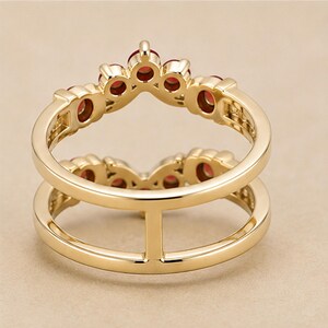 May include: A gold ring featuring a double band design connected by a vertical bar. The upper band is shaped like a crown and is set with five small, round, deep red gemstones. The metal has a polished, smooth finish throughout the entire piece.