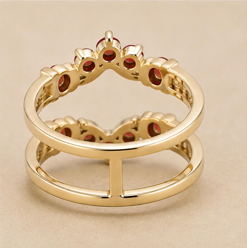 May include: A gold ring featuring a double band design connected by a vertical bar. The upper band is shaped like a crown and is set with five small, round, deep red gemstones. The metal has a polished, smooth finish throughout the entire piece.
