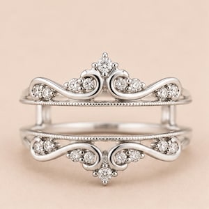 May include: A silver-toned ring guard featuring two curved bands connected by vertical bars. The design includes intricate scrollwork and milgrain detailing, accented with small, clear, round-cut stones set along the top and bottom edges.