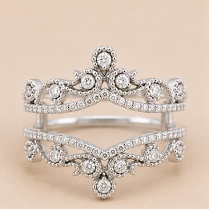May include: A silver-toned ring guard featuring two connected bands with a V-shaped design. The metal is embellished with numerous small, clear, round-cut stones and intricate beaded detailing along the edges of the metal framework.