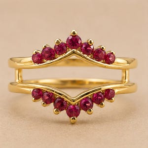 May include: A gold ring guard featuring two chevron-shaped bands connected by vertical bars. The top band is set with seven round, deep red rubies, and the bottom band is set with seven matching round, deep red rubies in a prong setting.