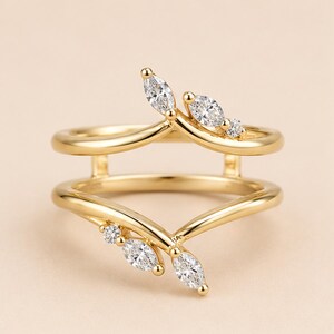 May include: A gold double-band ring featuring a V-shaped design on both bands. The top band is adorned with three marquise-cut clear stones, while the bottom band features three matching marquise-cut clear stones arranged in a leaf-like pattern.