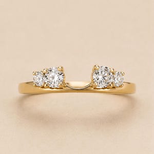 May include: A gold ring featuring a curved band design with two clusters of two round clear stones on each side. The stones are set in a prong setting, creating an open space in the centre of the band. The metal has a polished finish.
