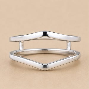 Peut inclure: Une bague en métal argenté poli présentant un design à double anneau avec une silhouette en forme de V. Les deux bandes sont reliées par de petites barres verticales, créant un espace ouvert entre les sections supérieure et inférieure.