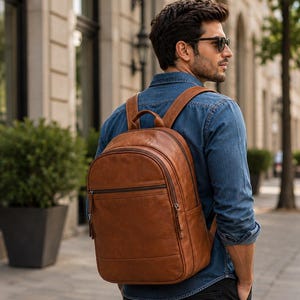 Può includere: Uno zaino in pelle color cuoio dotato di un ampio scomparto principale con chiusura a zip, una tasca frontale più piccola con zip, due spallacci regolabili e una maniglia superiore. La borsa presenta una texture liscia e cuciture visibili.