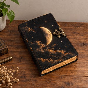 May include: A rectangular journal with a black cover featuring a crescent moon and clouds against a starry night sky. The journal has a metallic bronze-colored latch closure on the right side and thick, textured, light brown paper pages visible.