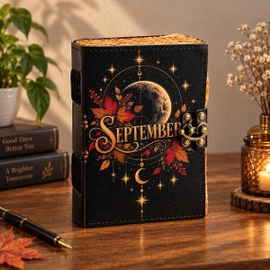 Pode incluir: Um diário preto com fecho de metal, apresentando um gráfico em tons dourados de uma lua, estrelas e folhas de outono. A palavra SEPTEMBER está impressa numa fonte decorativa no centro. O diário possui bordas texturizadas e uma lombada.