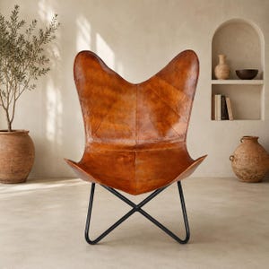 May include: A butterfly chair featuring a tan leather seat with visible stitching and a textured finish, supported by a black metal frame with a cross-base design. The chair has a high, curved backrest and a wide, deep seat for seating.