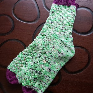 May include: A green and white patterned sock with a purple cuff. The sock is knitted with a textured stitch.