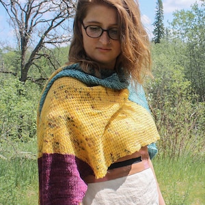 May include: A woman wearing a multi-colored knitted shawl with a yellow, teal, and purple color scheme. The shawl is draped over her shoulders and is made of a soft, textured yarn.