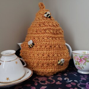 Puede incluir: Un calienta-tetera de ganchillo con forma de panal de abejas con pequeñas figuritas de abejas adheridas. El calienta-tetera es de color naranja cálido y está sobre una mesa con una taza de té y un platillo blancos y una taza de té floral.