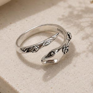May include: A silver-toned open ring featuring a snake design with embossed rose and leaf details along the band. The metal surface has an oxidised finish to highlight the intricate textures. The interior of the band is stamped with the text S925.