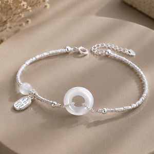 May include: A silver chain bracelet featuring a central white circular stone bead, a smaller round white bead, and a rectangular silver charm with engraved characters. The bracelet includes a delicate silver chain with a lobster claw clasp.