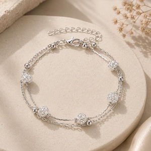 May include: A double-strand silver-toned bracelet featuring alternating smooth metallic beads and spherical charms encrusted with clear crystals. The piece includes a delicate chain link design and a secure lobster claw clasp closure.