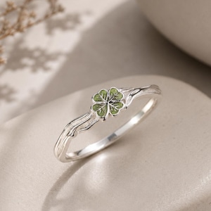 May include: A silver ring featuring a four-leaf clover motif with light green enamel detailing. The band has a textured, organic appearance that mimics the look of a thin, winding branch, leading up to the clover centerpiece at the top.