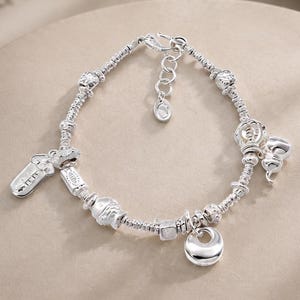 May include: A silver charm bracelet featuring a variety of small, polished silver charms including a fish, a rectangular tag, and circular shapes. The bracelet is constructed with a series of small silver beads and includes a lobster claw clasp.