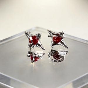 May include: A pair of silver-toned stud earrings featuring a four-pointed star shape with a smooth, polished finish. Each earring centres a small, round, deep red bead held in place by a thin, curved silver band that wraps around the star's centre.