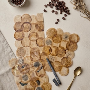 Può includere: Due fogli di carta sovrapposti con macchie circolari di caffè, caratterizzati da vari motivi a inchiostro nero, accenti in foglia d'oro e scarabocchi. Un pennarello grigio, un cucchiaio color ottone, una ciotola di caffè e fiori secchi.