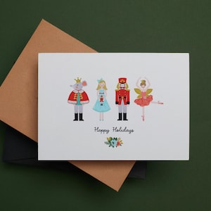 Peut inclure: Carte de vœux blanche avec des illustrations de personnages du ballet Casse-Noisette, dont un roi des souris, une fille, un casse-noisette et une ballerine. La carte indique "Joyeuses Fêtes" avec un motif floral. La carte est sur une enveloppe marron et noire.