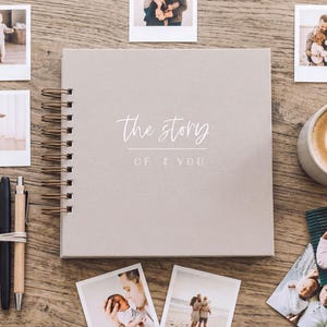 May include: A square, light grey spiral-bound photo album with the text "the story of you" printed on the cover. The album is surrounded by several square photographs of a family, a wooden baby rattle, three pens, and a ceramic mug of latte.