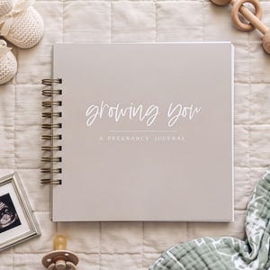 Growing You Pregnancy Journal with almond color cover laying on a bed surrounded by baby items