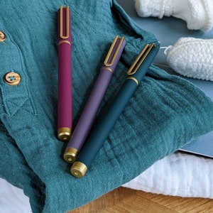 Peut inclure: Trois stylos avec des accents dorés et des corps de couleurs différentes : bordeaux, violet et sarcelle. Les stylos sont posés sur un tissu bleu et blanc.
