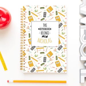May include: A spiral-bound notebook with a school supplies pattern and the text "This Notebook Belongs to Nicholas." Also, a red apple, yellow measuring tape, two pencils, and a white "AWESOME" sign.