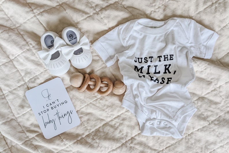 "I can't stop buying baby things" pregnancy milestone card next to baby shoes, rattle and baby onesie