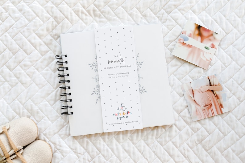 Puede incluir: Un diario de embarazo blanco con una encuadernaci&oacute;n en espiral negra. La portada presenta un dise&ntilde;o floral y el texto "The minimalist pregnancy journal 40 weeks of documenting the joys of your journey". La contraportada presenta un logotipo colorido para "nuts & bolts paper co."