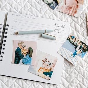 May include: An open white spiral bound journal with a black spine. The journal has two pages open, one with the words "Special Moments to Remember" and the other with the words "New Milestones / Exciting Moments". There are several photos attached to the pages, including a family photo, a photo of a baby, and a photo of a couple.
