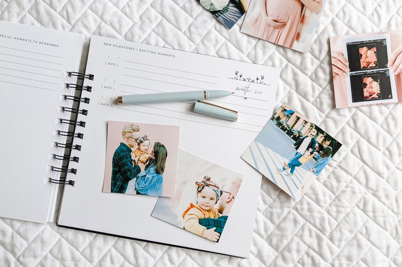 May include: An open white spiral bound journal with a black spine. The journal has two pages open, one with the words "Special Moments to Remember" and the other with the words "New Milestones / Exciting Moments". There are several photos attached to the pages, including a family photo, a photo of a baby, and a photo of a couple.