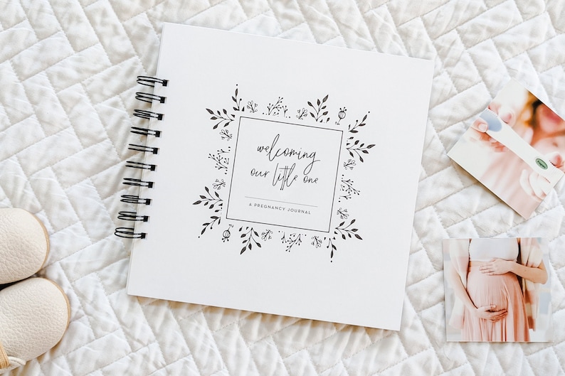 Puede incluir: Un diario de embarazo blanco en espiral con un dise&ntilde;o floral negro y el texto "welcoming our little one A pregnancy journal".