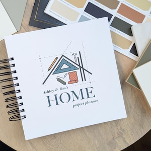 K&ouml;nnte beinhalten: Ein wei&szlig;es Spiralnotizbuch mit einer blauen und orangefarbenen Hausillustration und dem Text "Ashley & Tim's HOME project planner".
