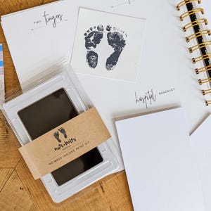 May include: A baby keepsake kit featuring a spiral-bound journal with sections for tiny fingers and hospital bracelets, a black inkless print pad with a brown paper band labeled nuts+bolts, a baby footprint card, and a photograph of a newborn.