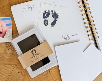 Puede incluir: Un kit de recuerdos para bebés que incluye un diario de espiral con secciones para dedos pequeños y pulseras de hospital, una almohadilla de tinta negra sin tinta con una banda de papel marrón etiquetada nuts+bolts, una tarjeta de huella y una foto.