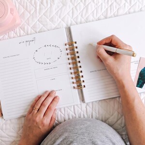 K&ouml;nnte beinhalten: Ein spiralgebundenes Schwangerschaftstagebuch mit wei&szlig;en Seiten, die gedruckte Abschnitte wie "we are pregnant", "how we found out" und "how we told others" enthalten. Eine hellrosa Sofortbildkamera und zwei quadratische Fotos liegen daneben.